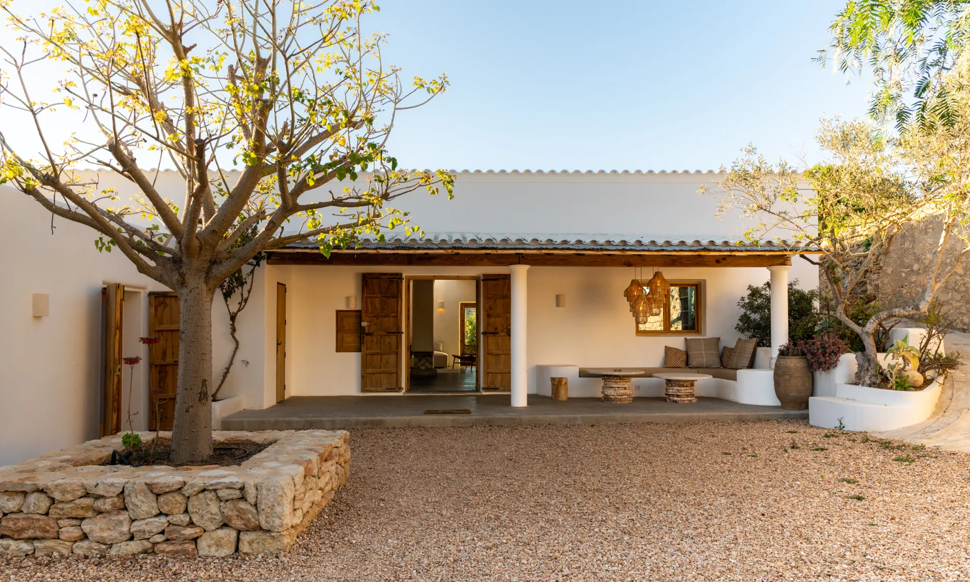 Outdoor patio of a white stucco house with wooden doors, stone planter with a tree, and built-in bench seating with cushions under hanging woven lamps.