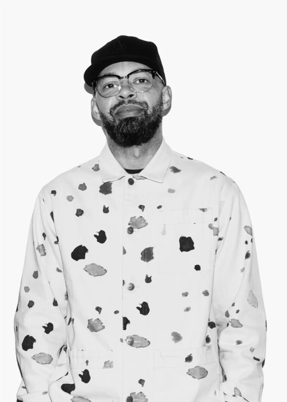 Kristian Smith with beard and glasses wearing a spotted jacket and a baseball cap against a white background.