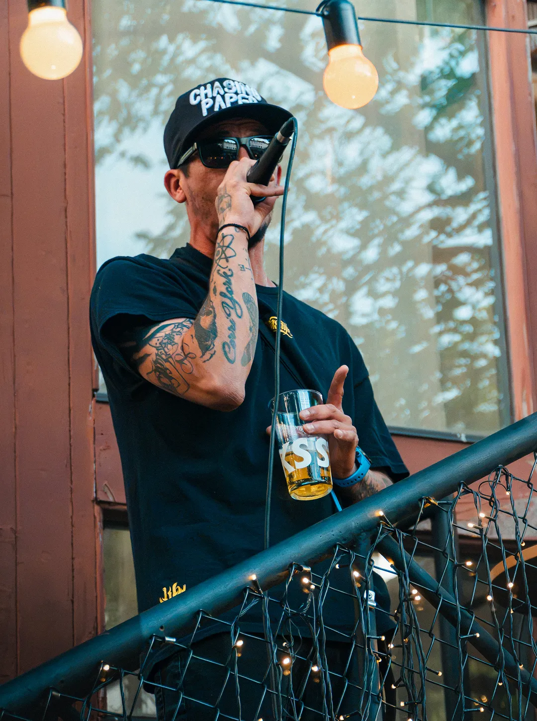 Man wearing sunglasses and a hat sings into a microphone holding a beer glass on decorated staircase.