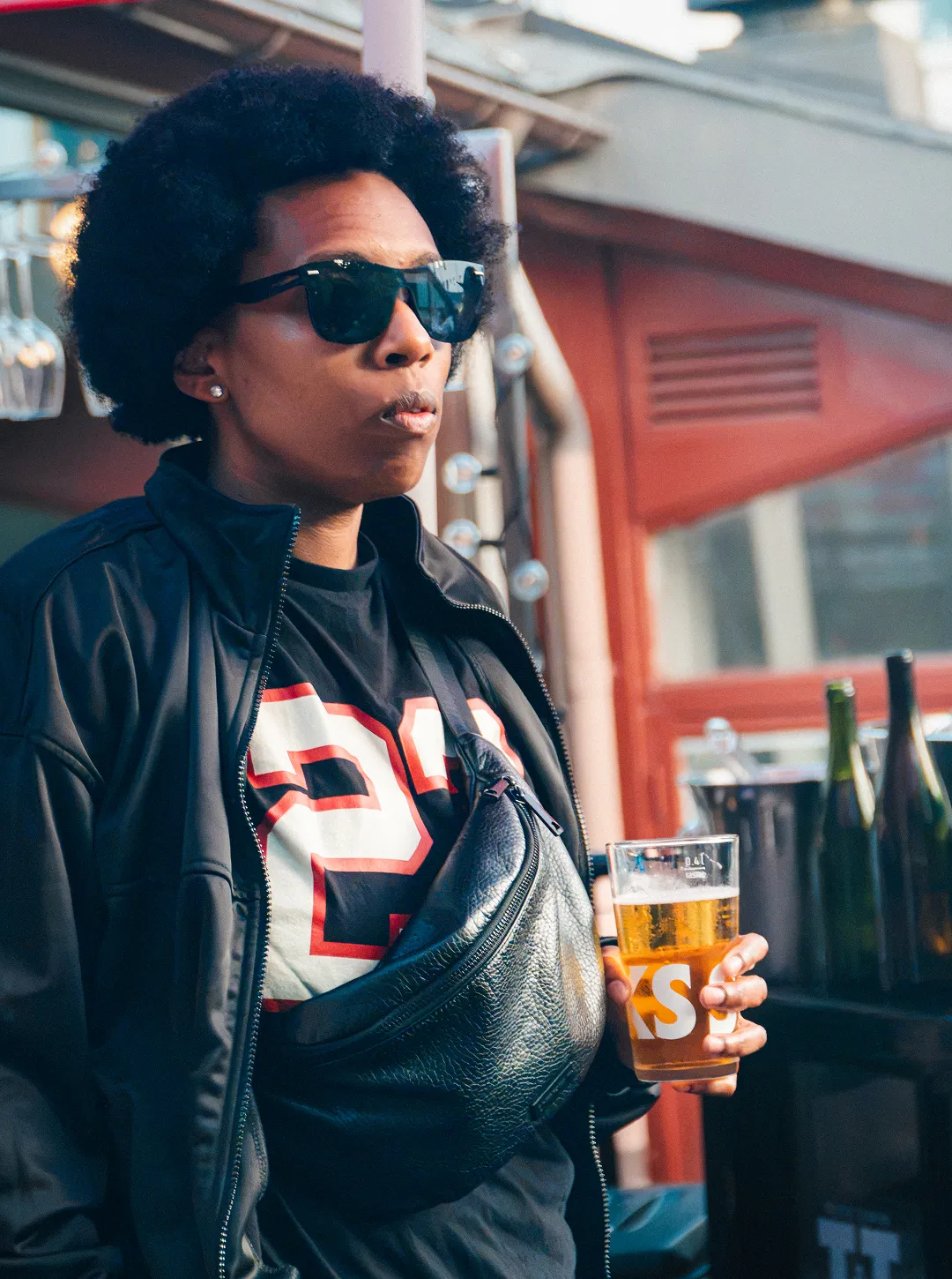Person wearing sunglasses and black jacket holding a glass of beer in an outdoor setting.
