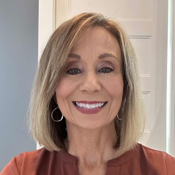 Smiling middle-aged woman with shoulder-length blonde hair wearing a rust-colored top and hoop earrings.