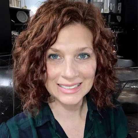 Woman with curly red hair and blue eyes smiling, wearing a dark green and blue plaid shirt.