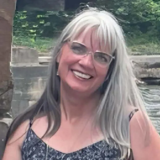 Smiling woman with long hair dyed half black and half white, wearing glasses and a patterned sleeveless top, standing outdoors near a stone structure and greenery.