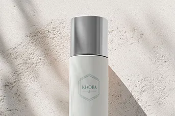 Close-up of a white cylindrical skincare bottle with a silver cap labeled 'KIAORA' on a textured light surface with shadows.