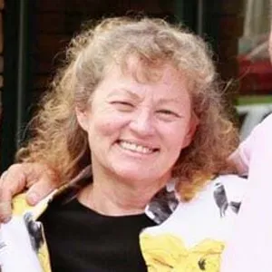 Smiling woman with curly light brown hair wearing a black top and a light-colored jacket with yellow floral details.