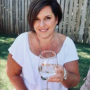 Smiling woman with short dark hair wearing a white blouse, holding a glass of white wine outdoors near a wooden fence.