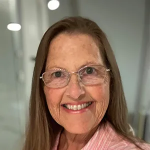 Smiling elderly woman with glasses and long brown hair wearing a pink top in a softly lit indoor setting.