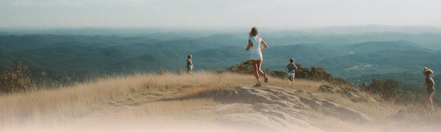 Three children and a woman running on a rocky hilltop with tall grass overlooking layered blue mountains under a hazy sky.