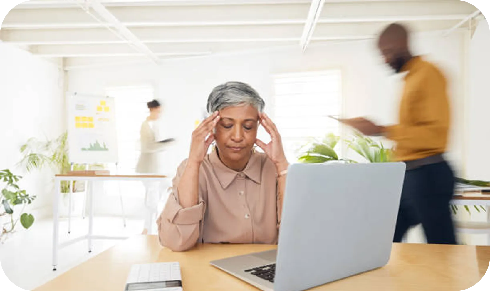 Woman sitting at a desk with a laptop, holding her temples with a stressed expression, while two blurred people walk in the background of a bright office.