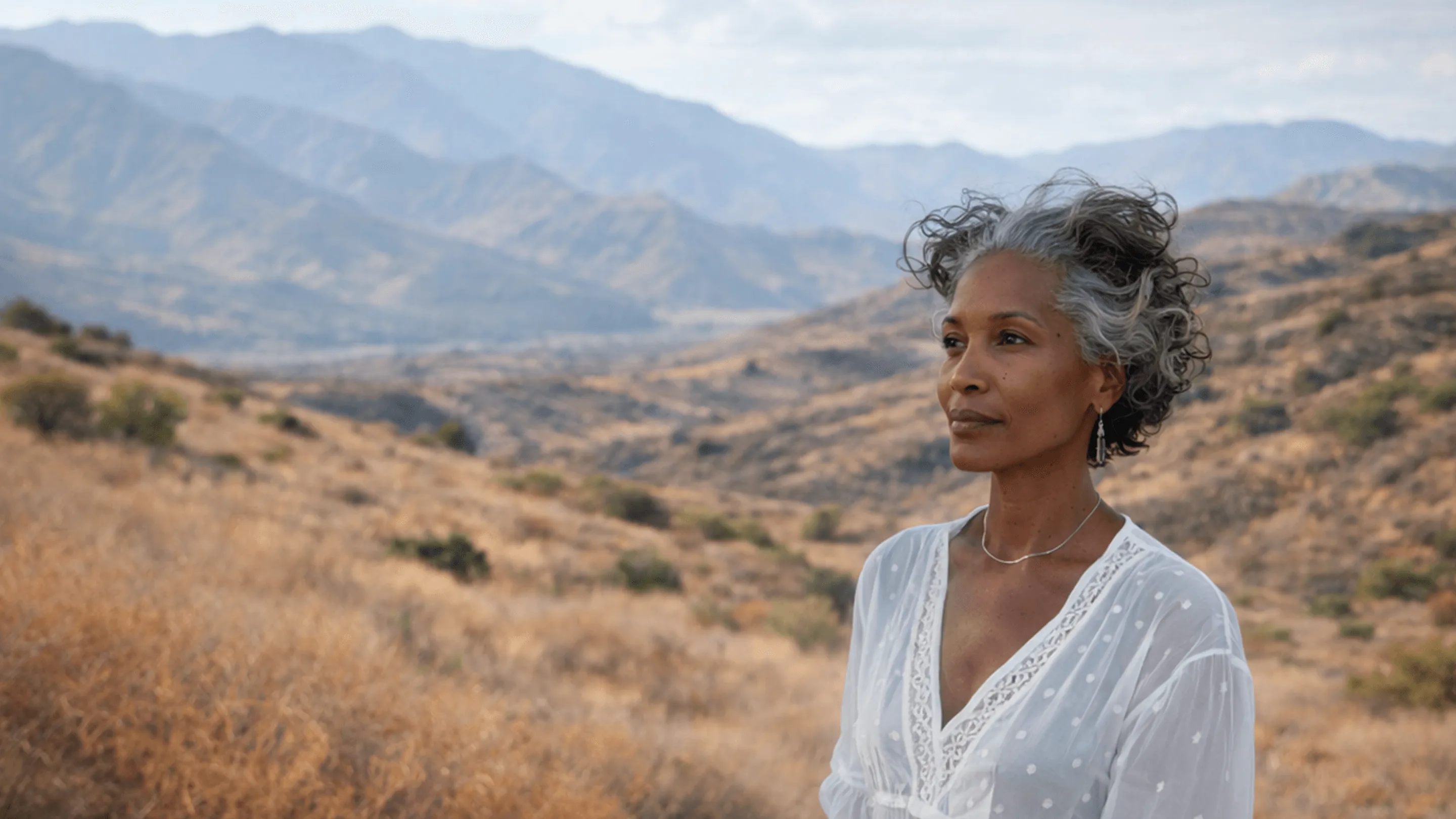 Middle-aged woman with curly gray hair wearing a white blouse standing in a dry mountainous landscape under a cloudy sky.