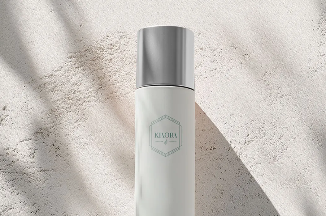 Sleek white cylindrical skincare bottle with silver cap labeled Kiaora, placed on textured beige surface with shadows.