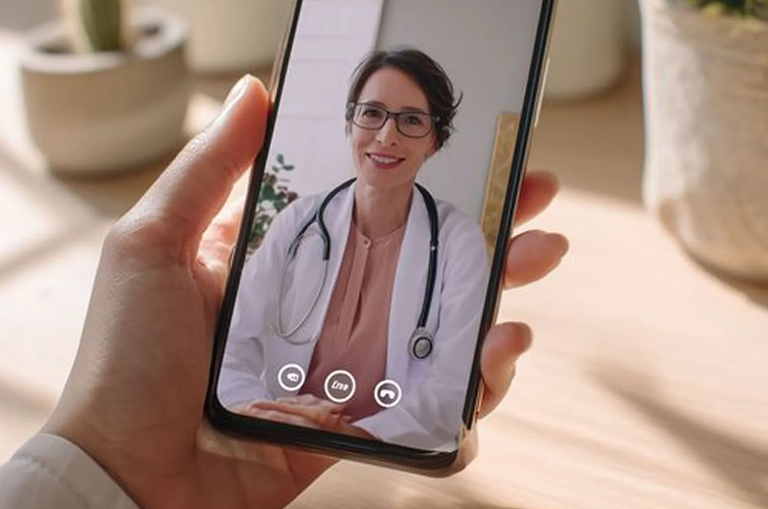 Hand holding a smartphone showing a video call with a smiling female doctor wearing glasses and a white coat with a stethoscope.