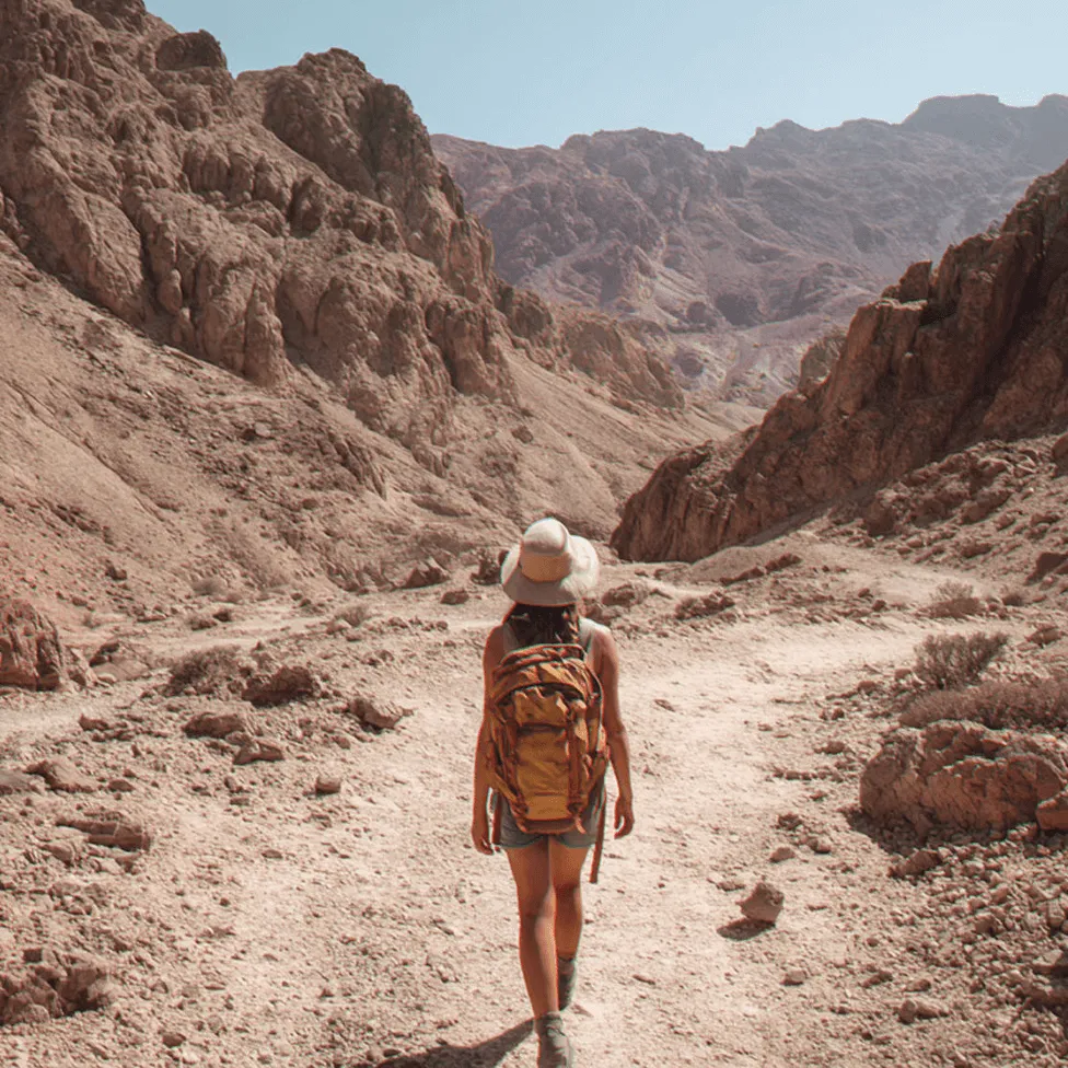 Person with a backpack and hat hiking on a rocky, mountainous trail under a clear sky.