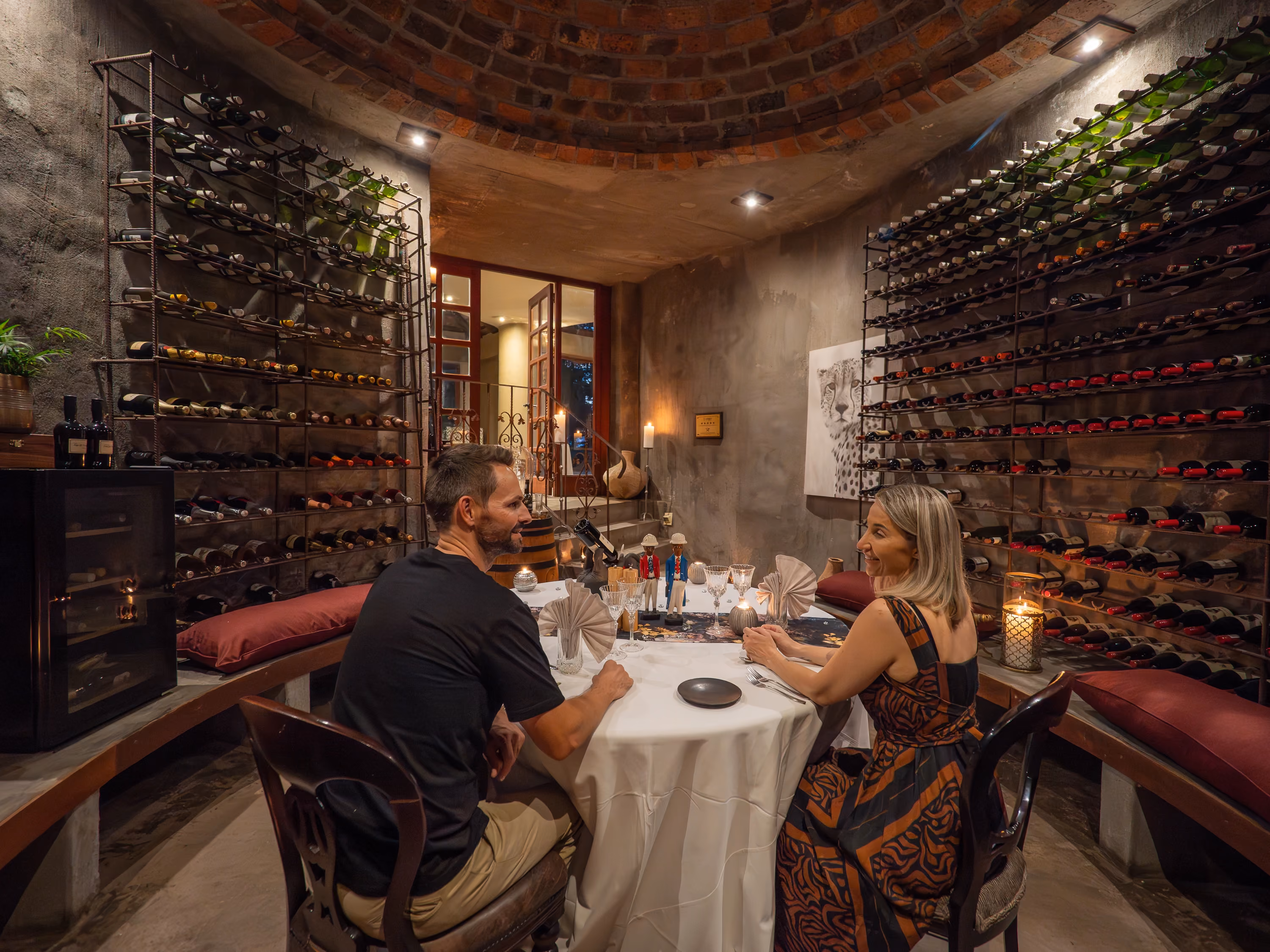 A couple smiling and enjoying an intimate dinner at an elegantly set table inside a rustic, stone-walled private wine cellar surrounded by curved brick ceilings and floor-to-ceiling iron wine racks.