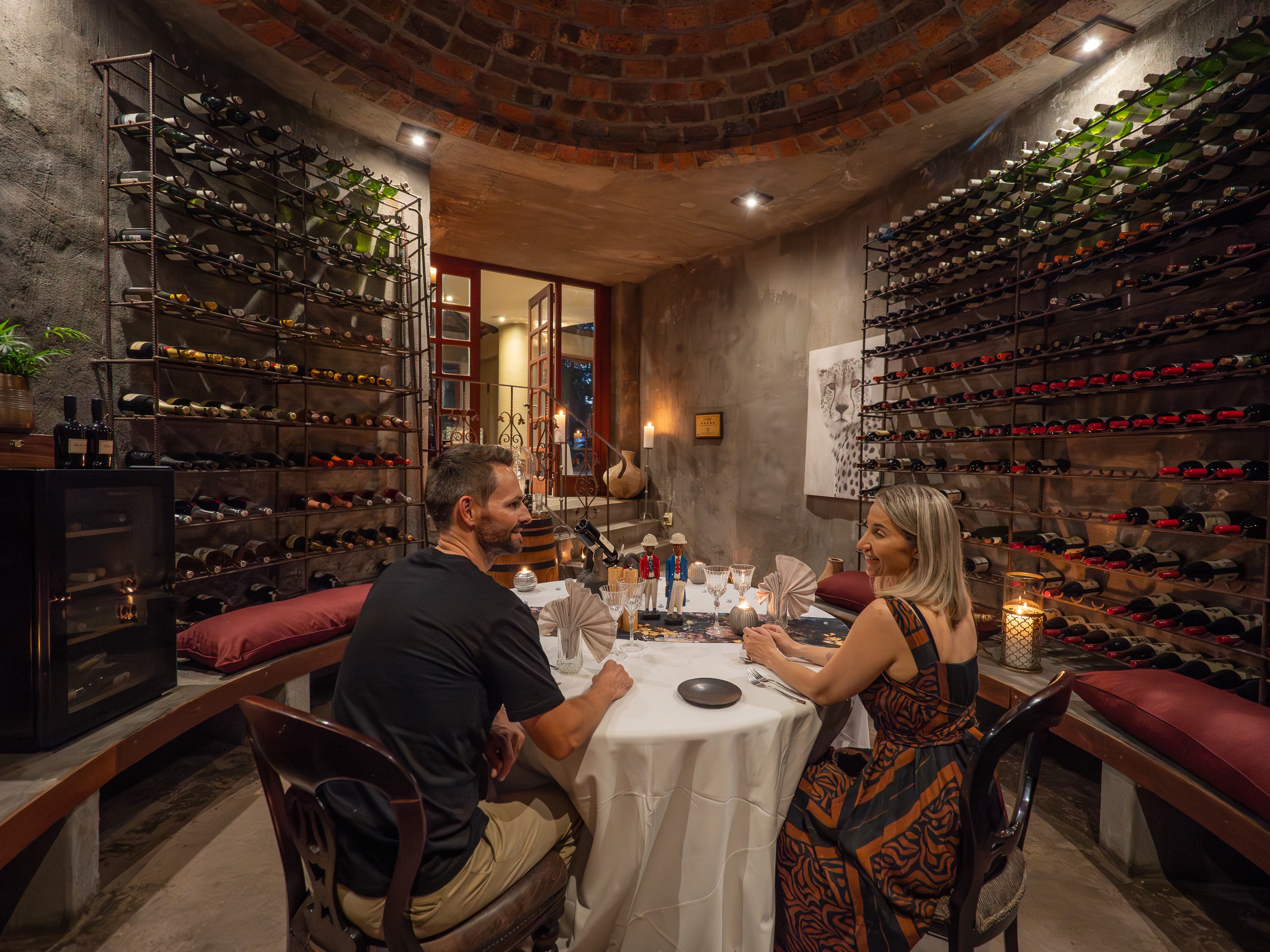 A couple smiling and enjoying an intimate dinner at an elegantly set table inside a rustic, stone-walled private wine cellar surrounded by curved brick ceilings and floor-to-ceiling iron wine racks.