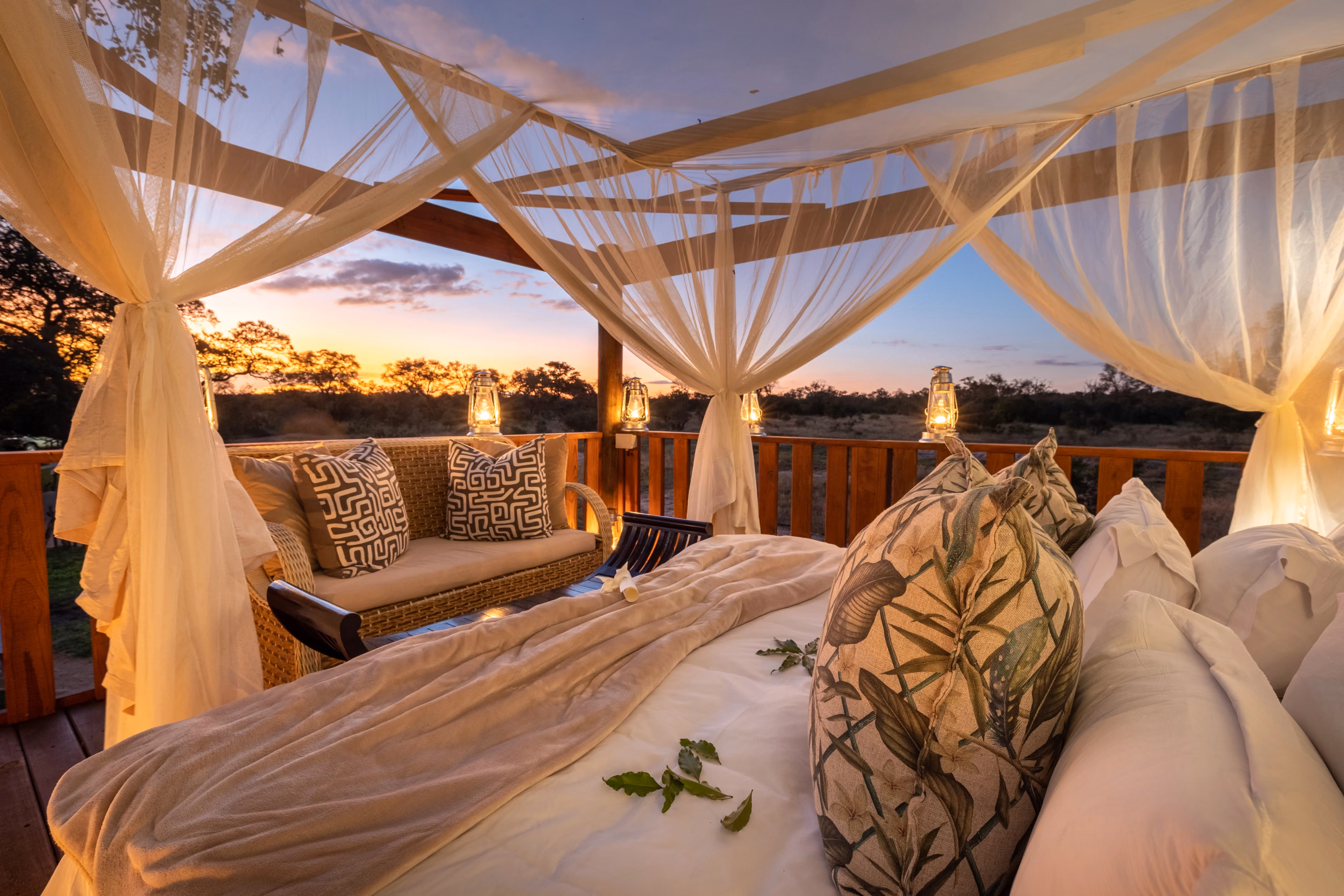 A luxury open-air sleepout deck at sunset, featuring a large bed with draped white mosquito netting, comfortable outdoor seating, and warm lanterns overlooking the African bush.