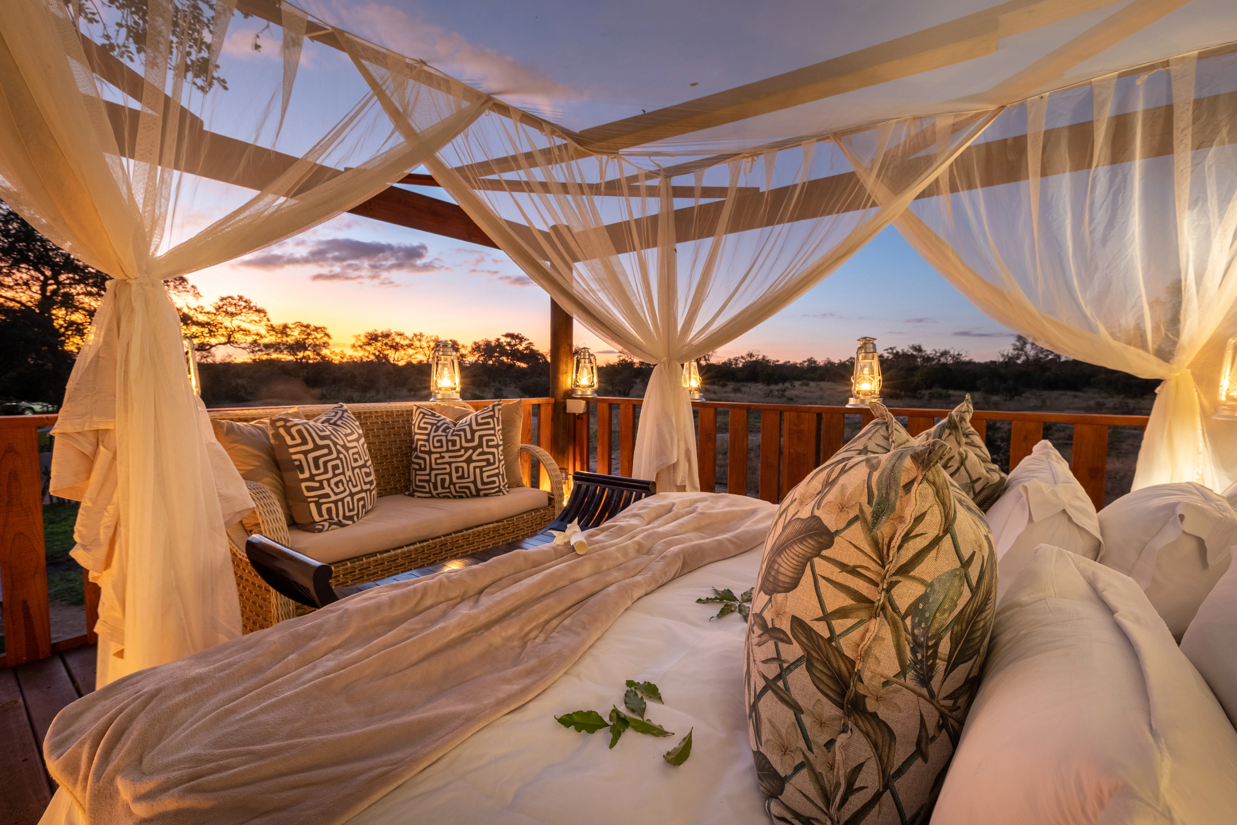 A luxury open-air sleepout deck at sunset, featuring a large bed with draped white mosquito netting, comfortable outdoor seating, and warm lanterns overlooking the African bush.