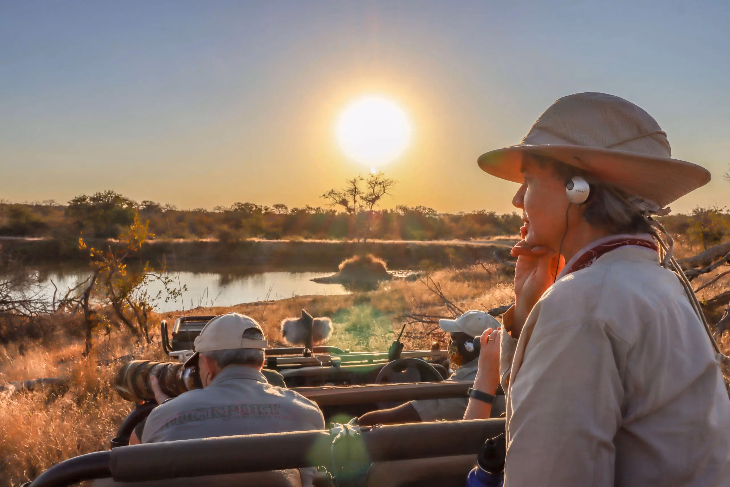 A safari guest in a wide-brimmed hat wearing an earpiece to listen to nature sounds from an open-top vehicle at sunrise, overlooking a tranquil waterhole in the bushveld.