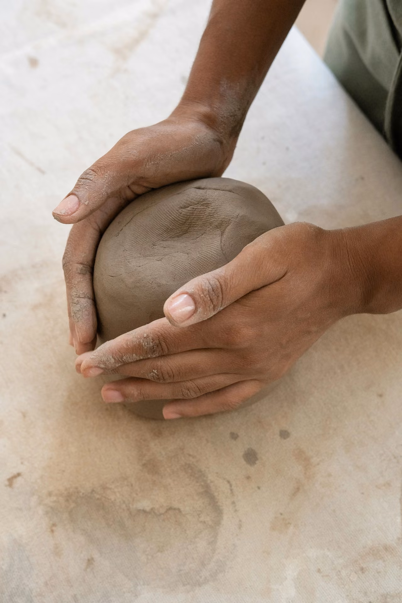 A close-up shot of an artisan's hands gently shaping and molding a smooth mound of natural clay on a pottery workshop table.