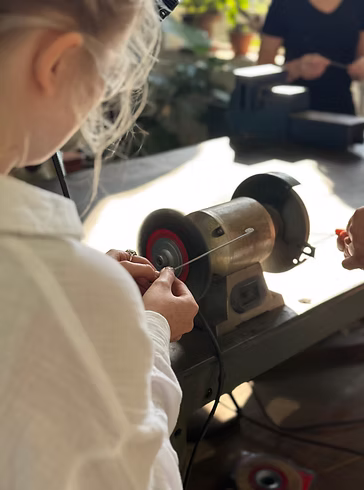 A workshop participant carefully crafting jewelry, using a bench grinder machine tool to smooth down a piece of metal wire at a naturally lit workbench.