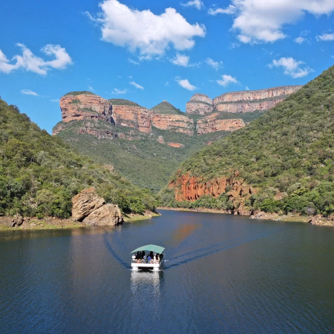 An aerial view of a white passenger boat cruising down a calm river surrounded by the massive, lush green mountain cliffs of the Blyde River Canyon under a bright blue sky.