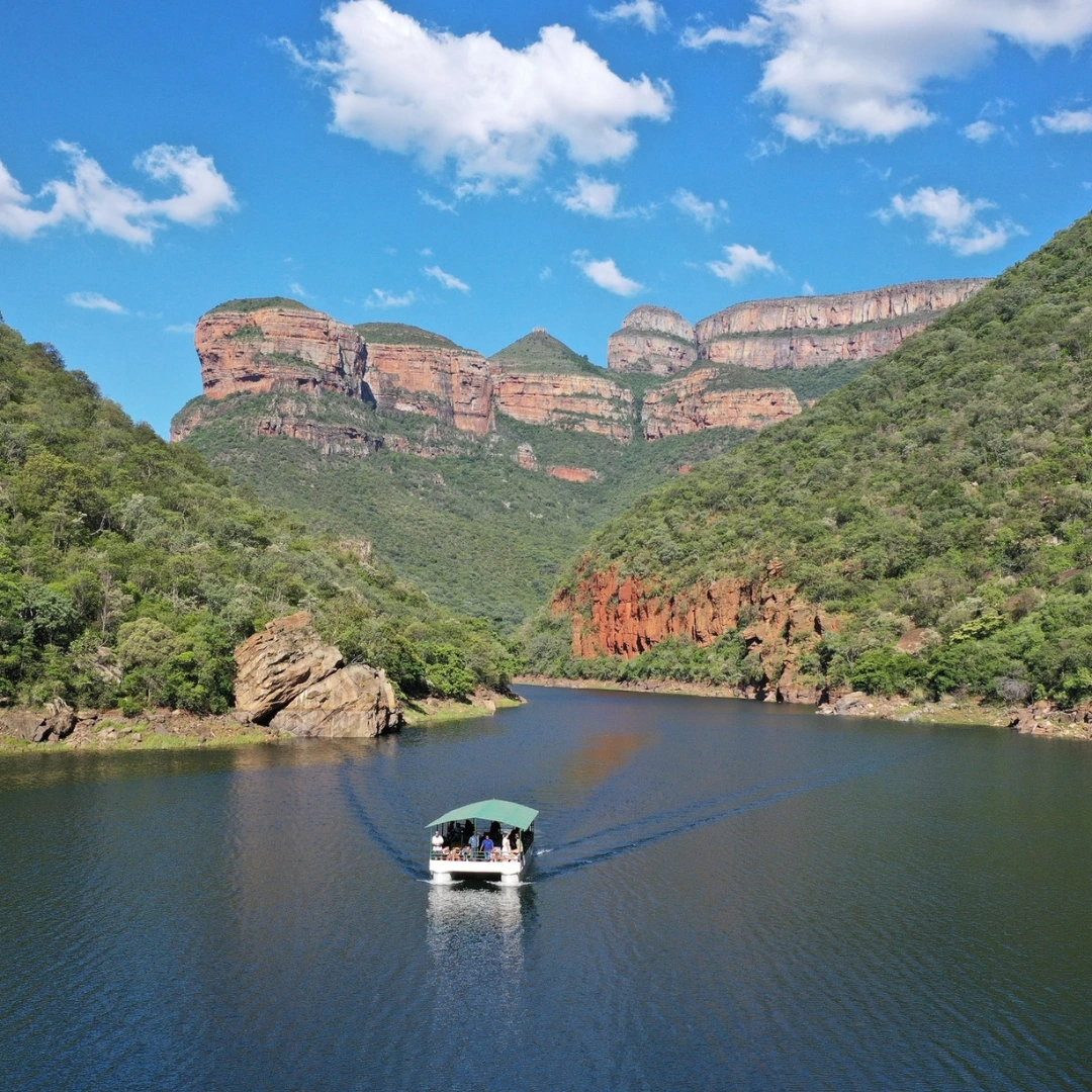 An aerial view of a white passenger boat cruising down a calm river surrounded by the massive, lush green mountain cliffs of the Blyde River Canyon under a bright blue sky.