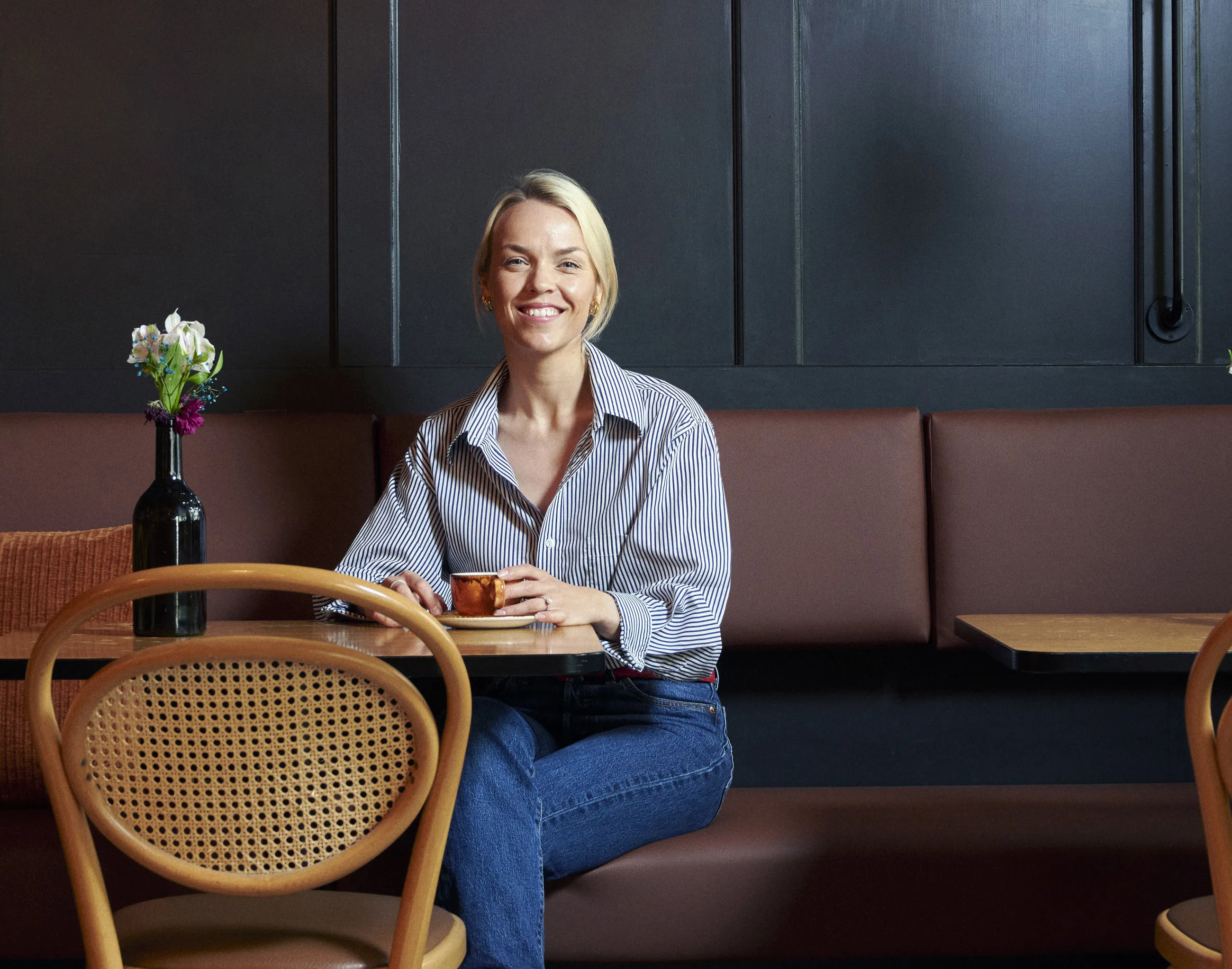 Kvinne som sitter og smiler på en benk i et vakkert lokal med en kopp varm drikke