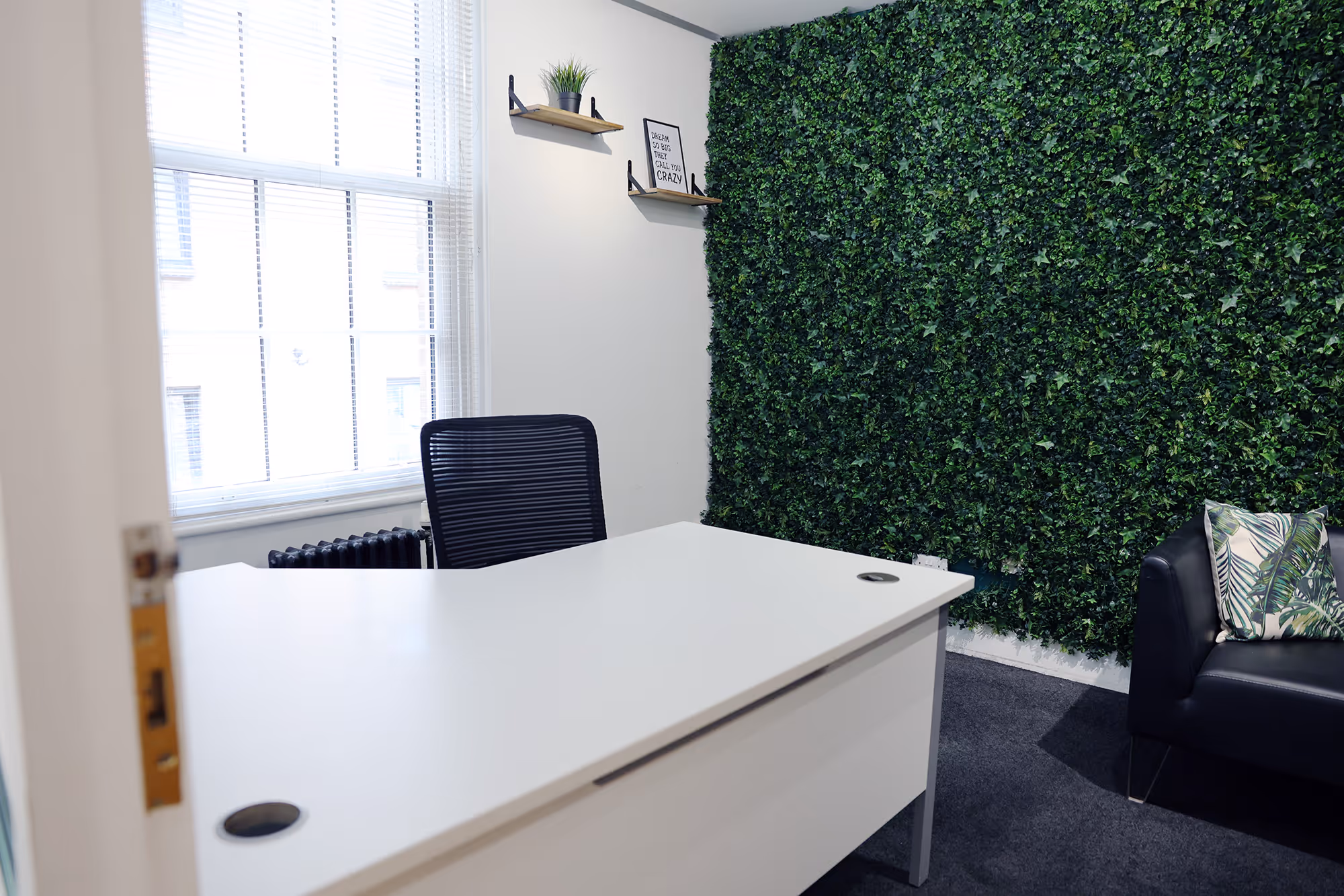 Minimalist office with white desk, black chair, green leaf wall, and black sofa with leaf-patterned pillow.