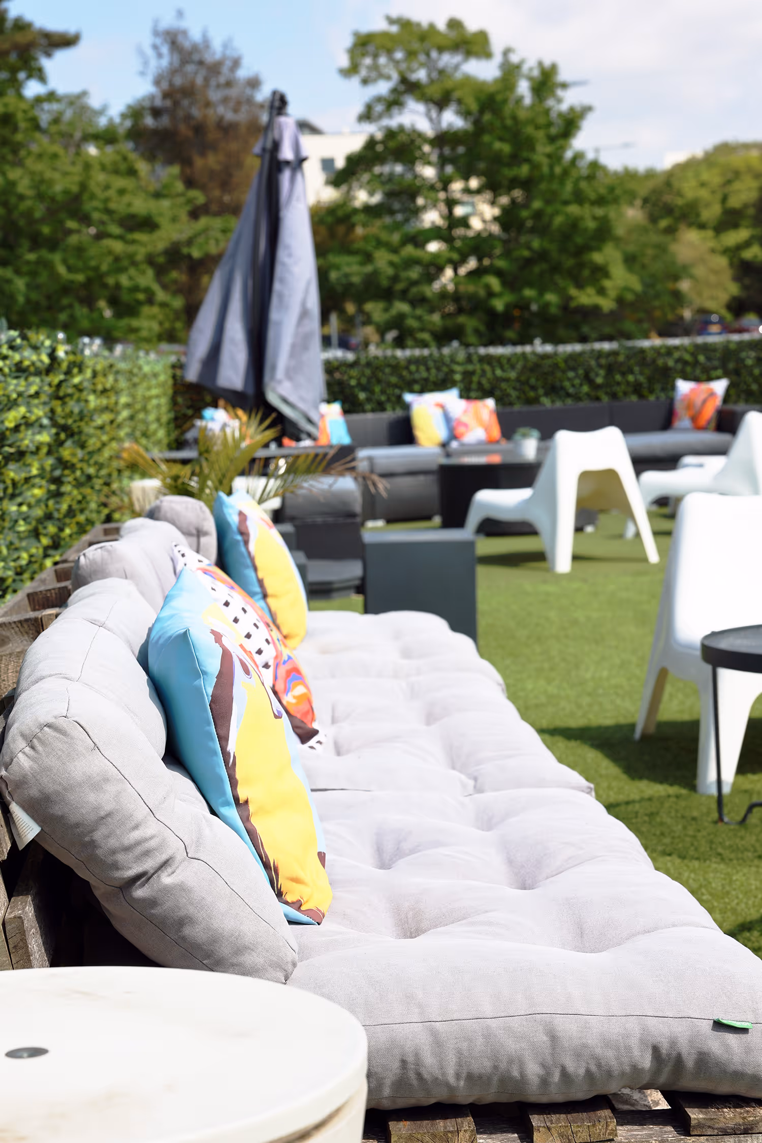 Outdoor seating area with cushioned benches, colorful pillows, white chairs, and a closed umbrella on green lawn.
