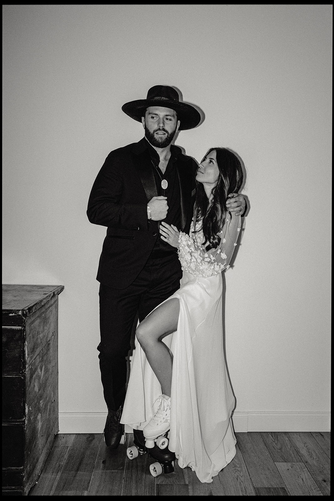 Black and white portrait of a couple posing in a room, the groom in a wide-brimmed hat