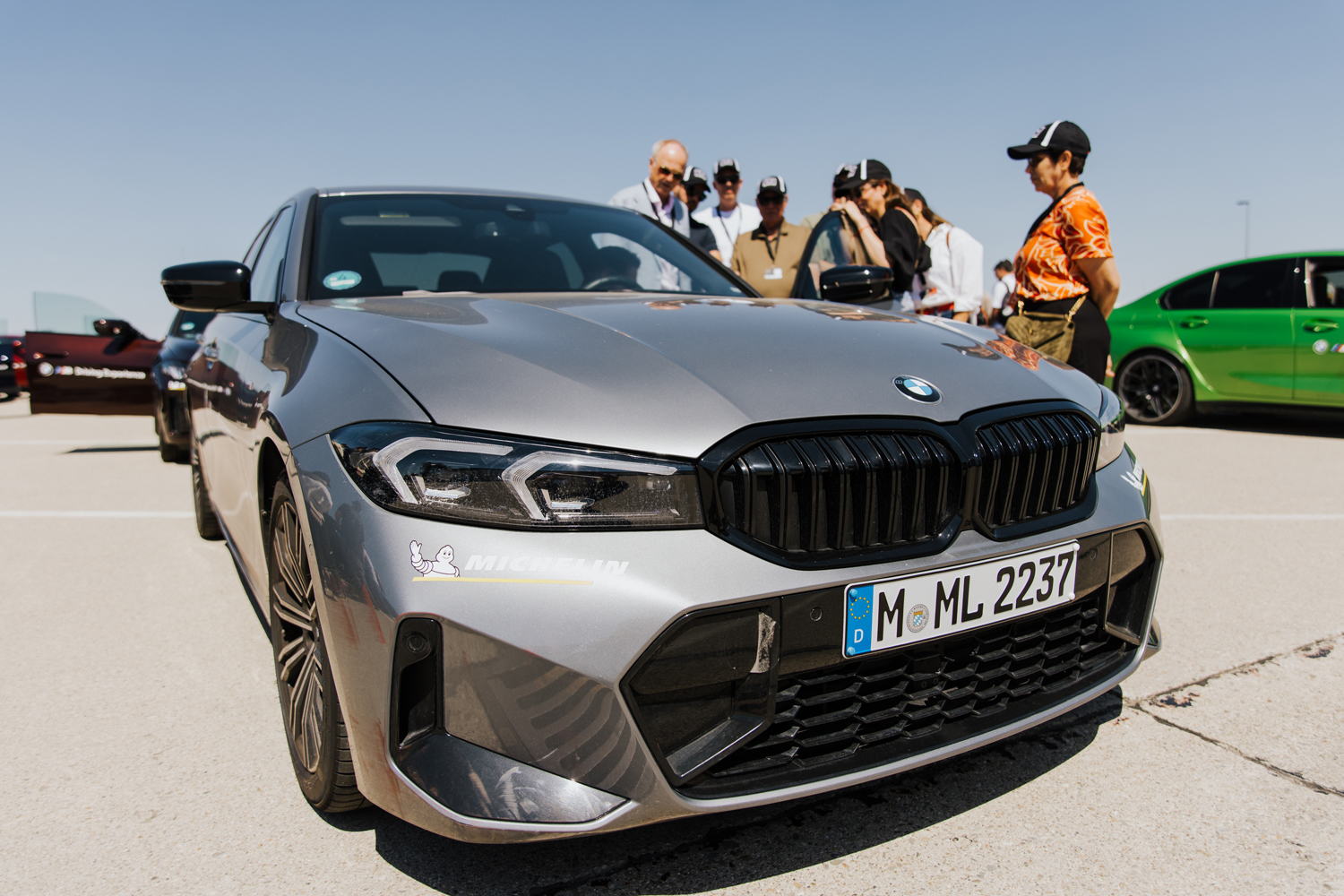 Detailaufnahme eines modernen grauen BMW vor einer Gruppe von Event-Teilnehmern unter blauem Himmel – professionelle Autofotografie in München.