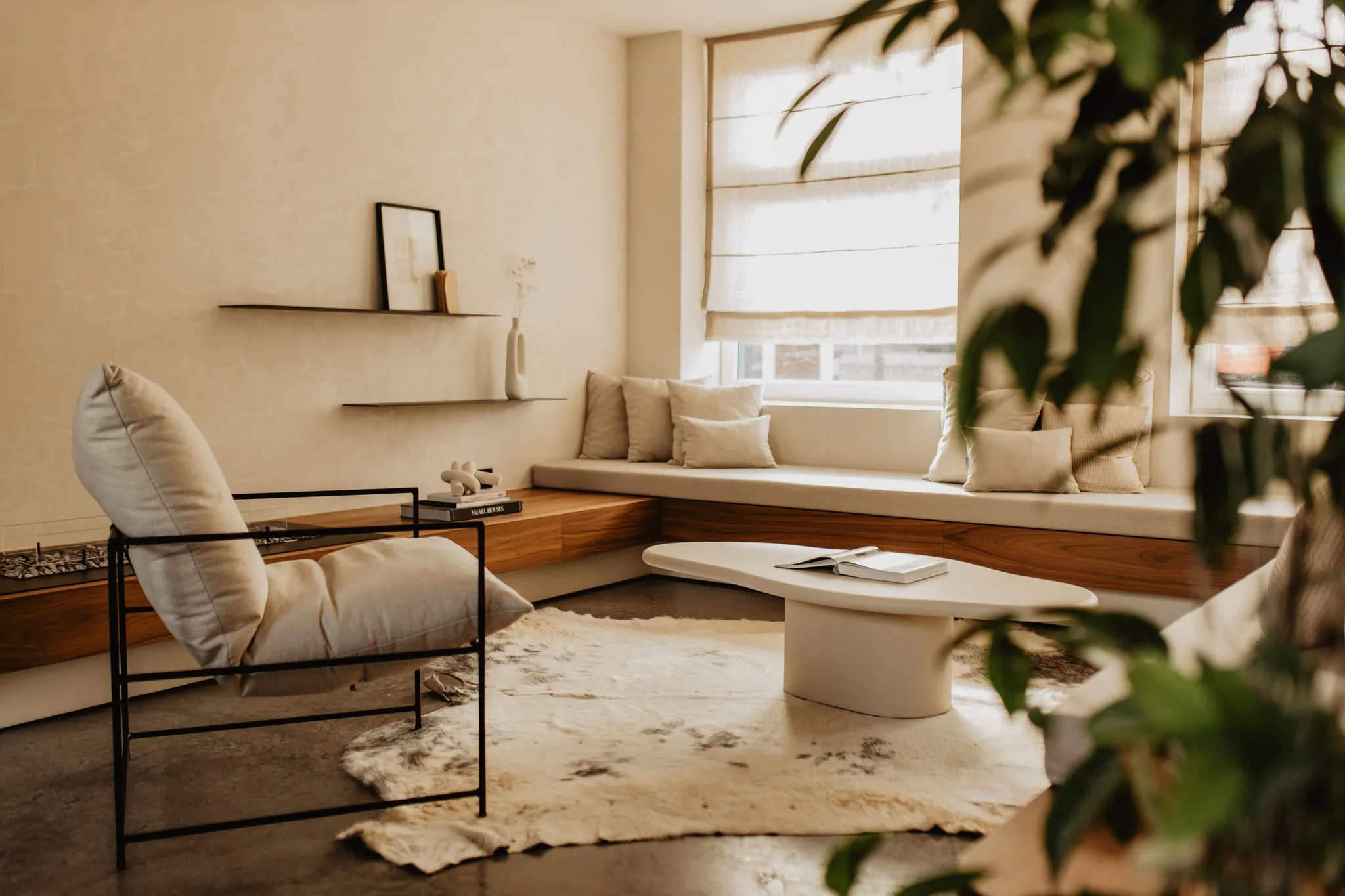 A modern living room with a built-in window seat adorned with cushions, a sleek coffee table, an armchair, and houseplants, creating a cozy atmosphere.