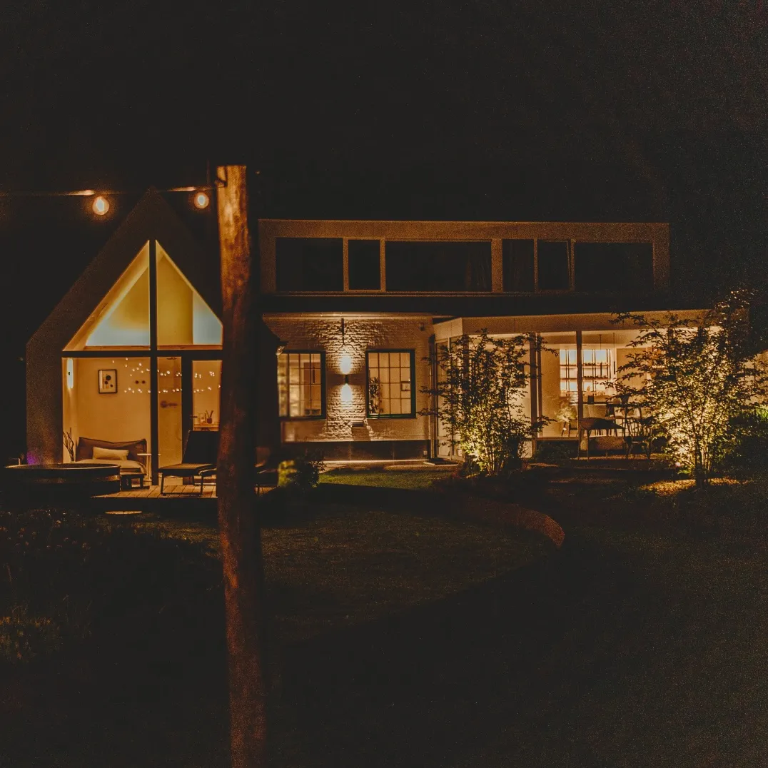 A warmly lit modern house at night, with visible interior lighting and exterior landscaped garden lights.