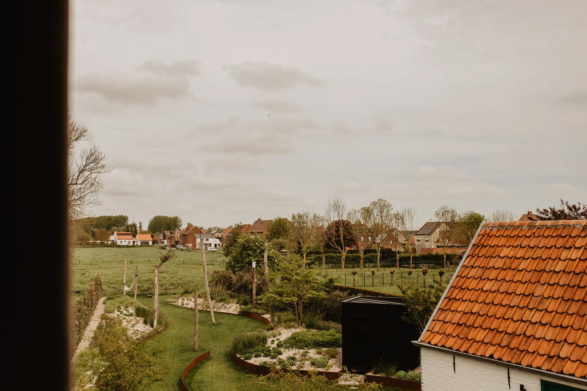 The image shows a rural landscape with houses, a well-kept garden, and fields under a cloudy sky, likely viewed from a window.