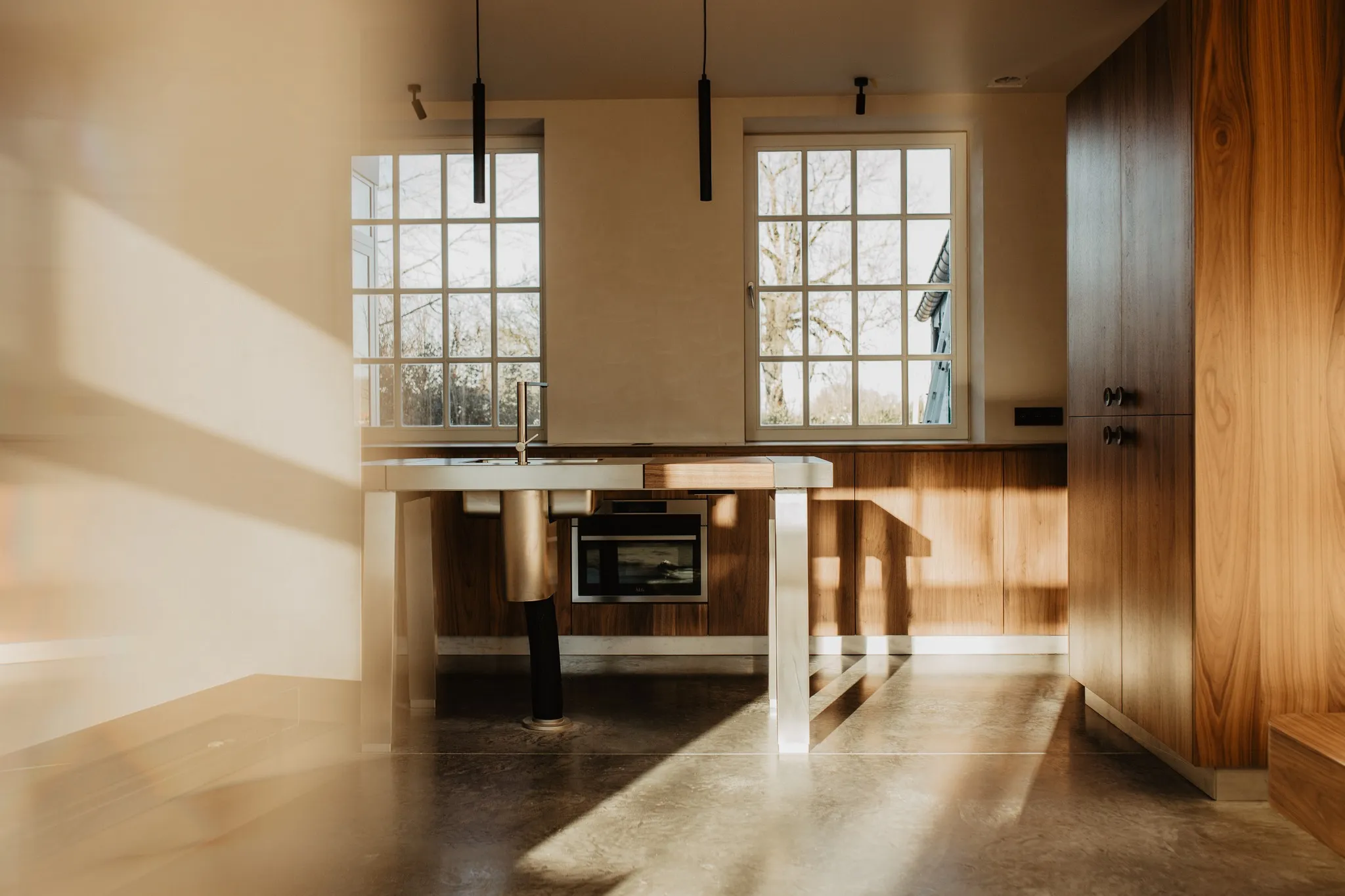 A modern kitchen with wooden cabinets, a central island with a built-in sink and oven, under natural light coming through large windows.
