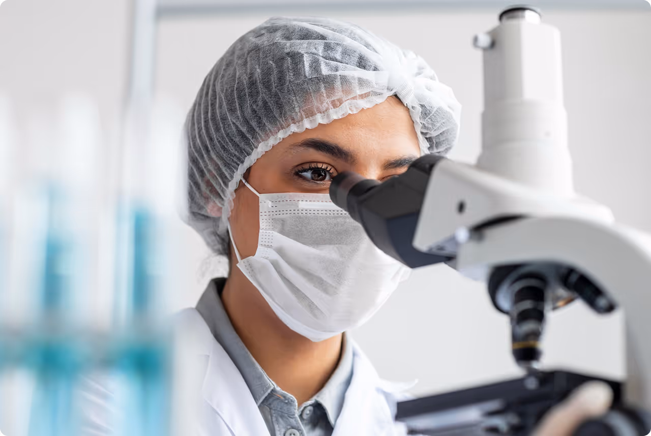 Pesquisadora com máscara e touca descartável olhando para o microscópio.