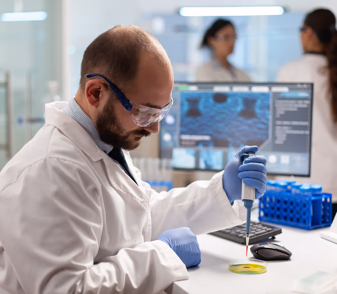Cientista de jaleco e luvas azuis usando pipeta para transferir líquido vermelho em uma placa de Petri em laboratório moderno.