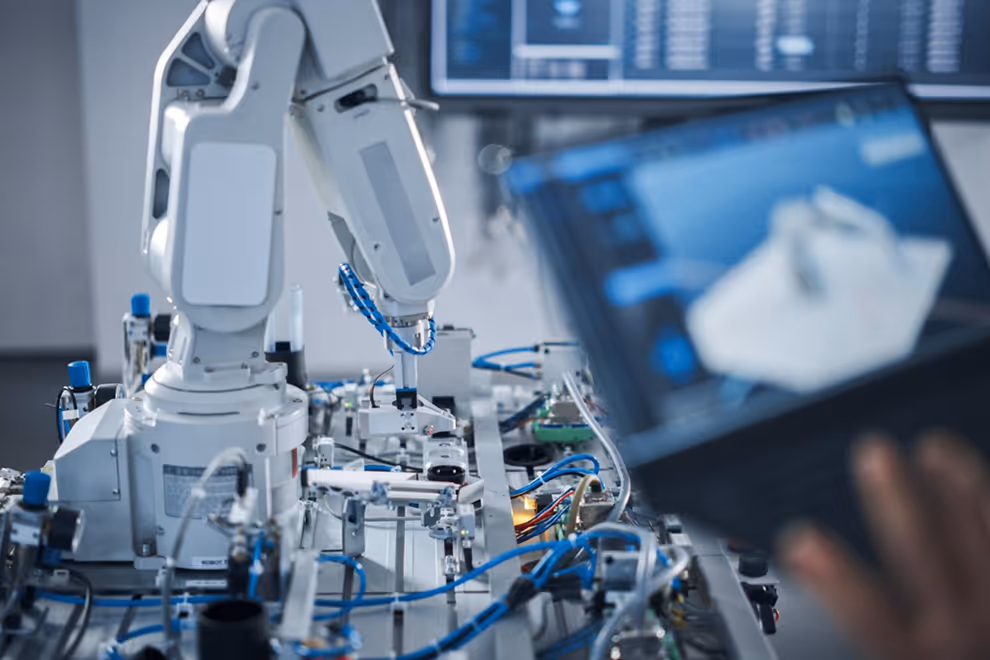 Braço robótico operando máquina industrial com controle em tela de computador ao fundo.