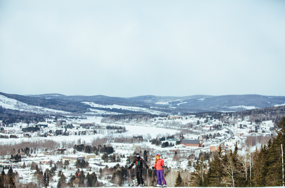two skiers pause at a ski mountain in fort kent with the st john river and catholic church far off in the distance. 