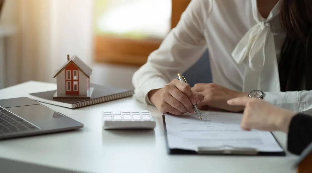 Woman with pen in hand, sitting at a desk to sign a document. On the desk sits a small house figurine.