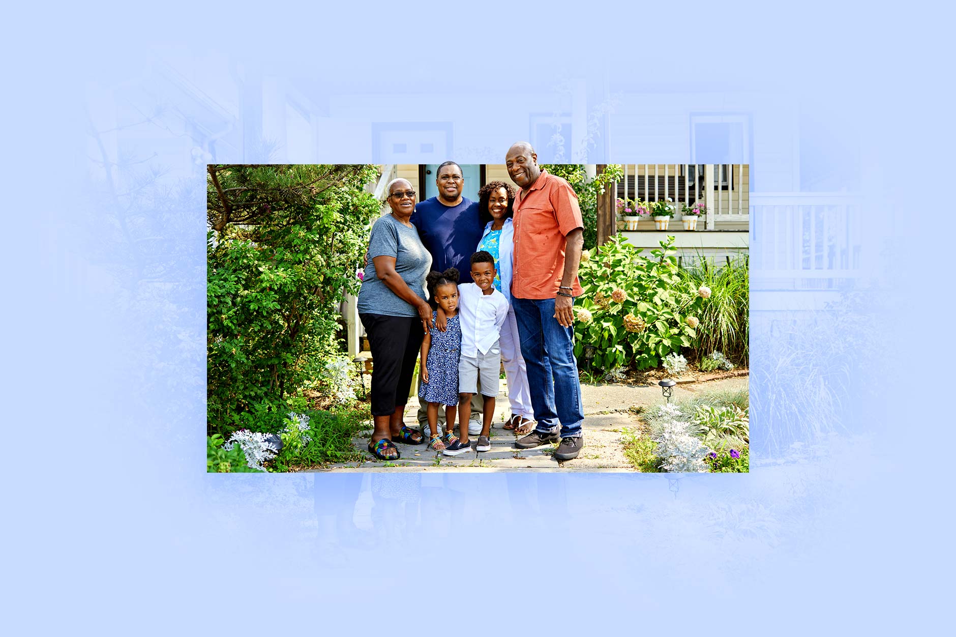 A multigenerational family stands together in front of a home with lush foliage and flowers.