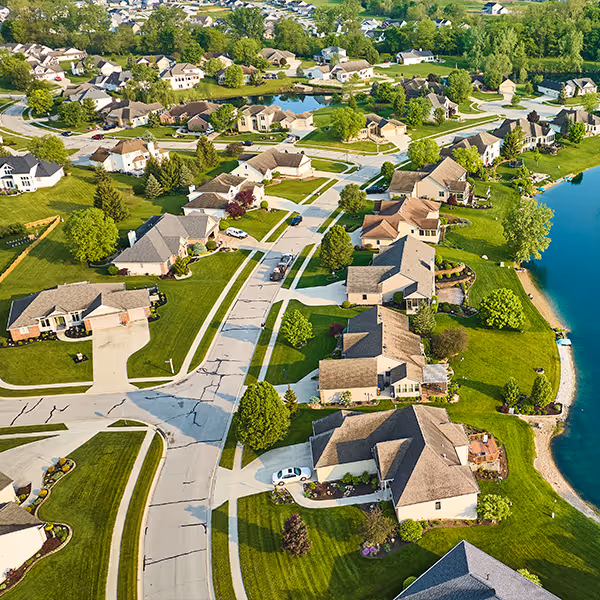 Aerial view of a suburban neighborhood with houses, green lawns, roads, trees, and a lake.