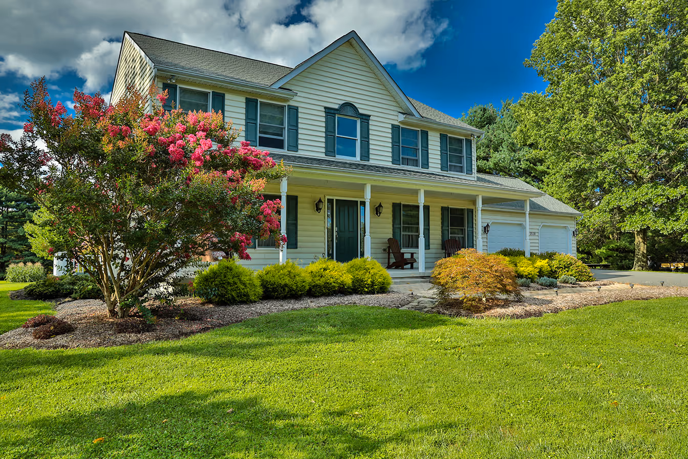 Two-story house with white siding, green shutters, porch, and well-kept lawn with colorful bushes.