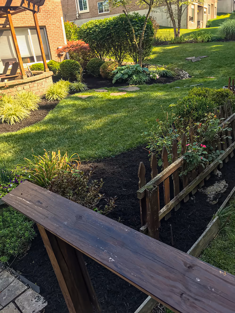 Well-maintained garden with green grass, shrubs, wooden fence, and a seating area near a brick house.