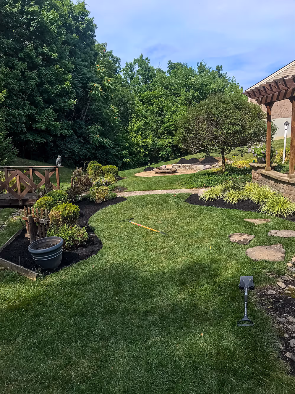 Well-maintained backyard with green lawn, garden beds, a small wooden bridge, and gardening tools on grass.