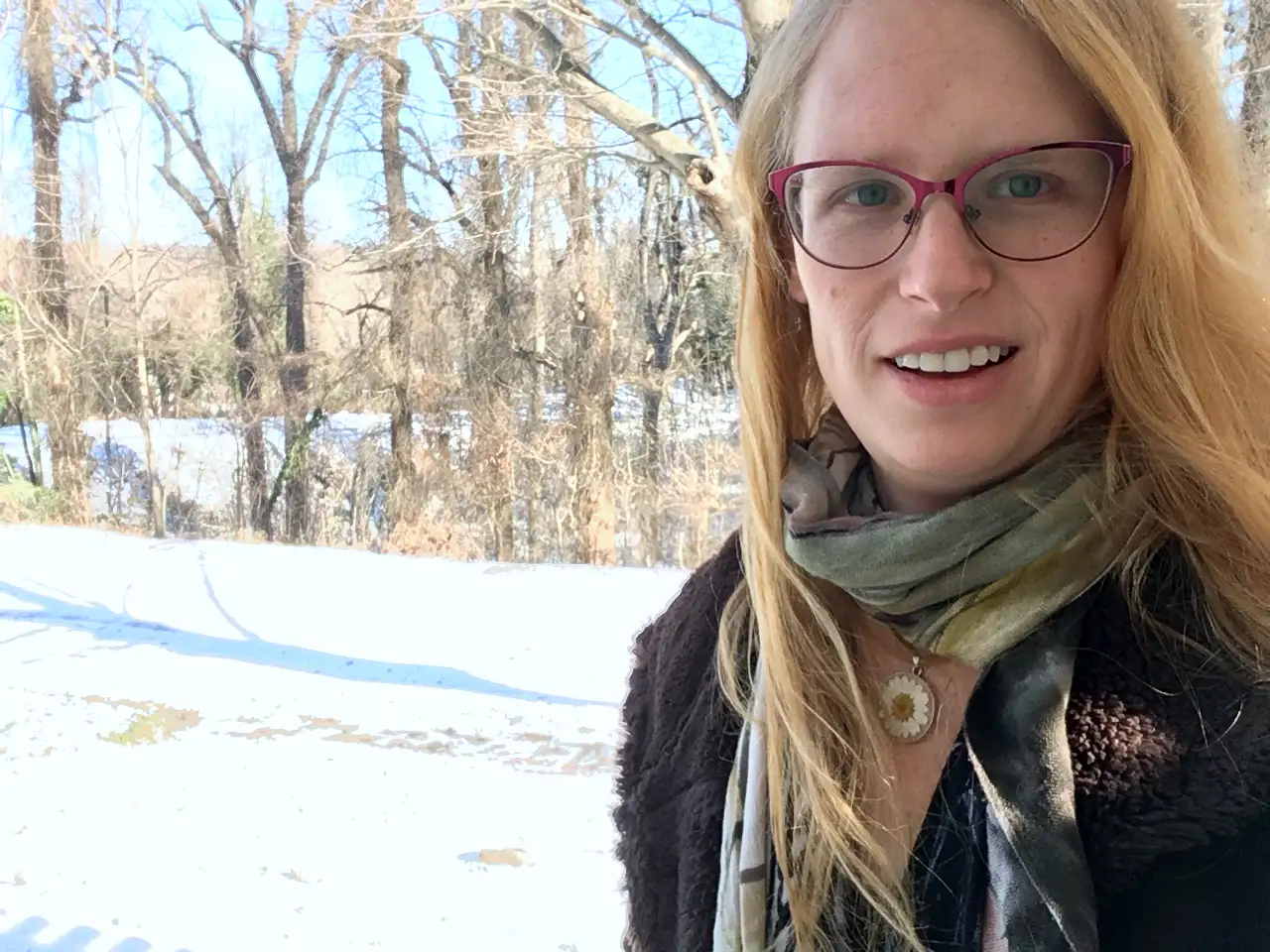 Evelyn standing on a snowy road with trees in the background. She is looking at the camera, her blond hair swept to the side, wearing a brown coat, a scarf, and a flower necklace.