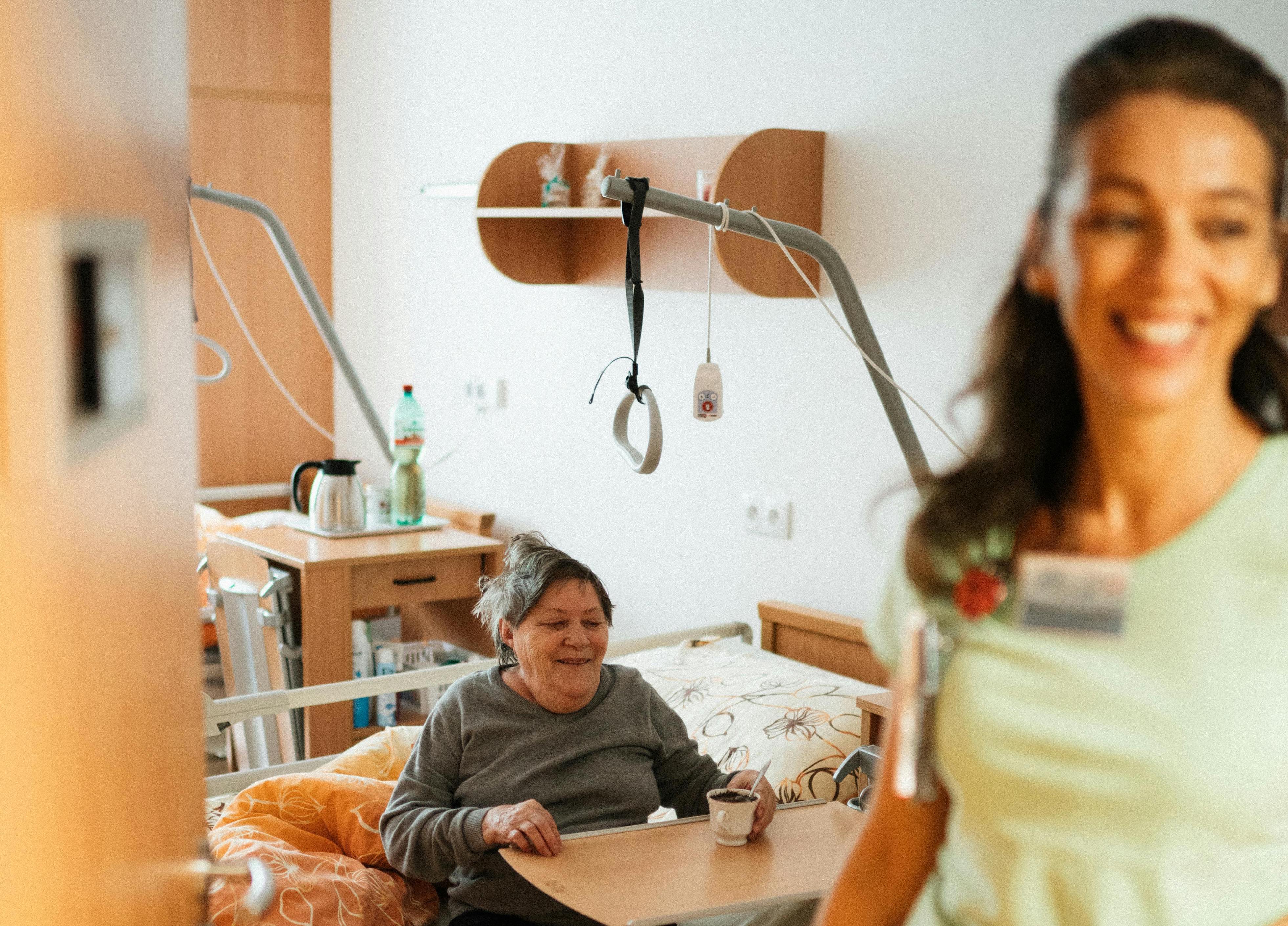 Anziana sorridente a letto in ospedale con una tazza ed una badante infermiera accanto.