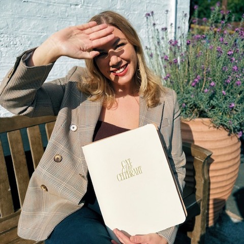 Cat the Celebrant, LGBTQ+ specialist wedding celebrant for engaged couples is holding a cream folder that says cat the celebrant. Cat is smiling and sits in the sun outside a wedding venue with her hand held up to her face to shield from the sunlight.