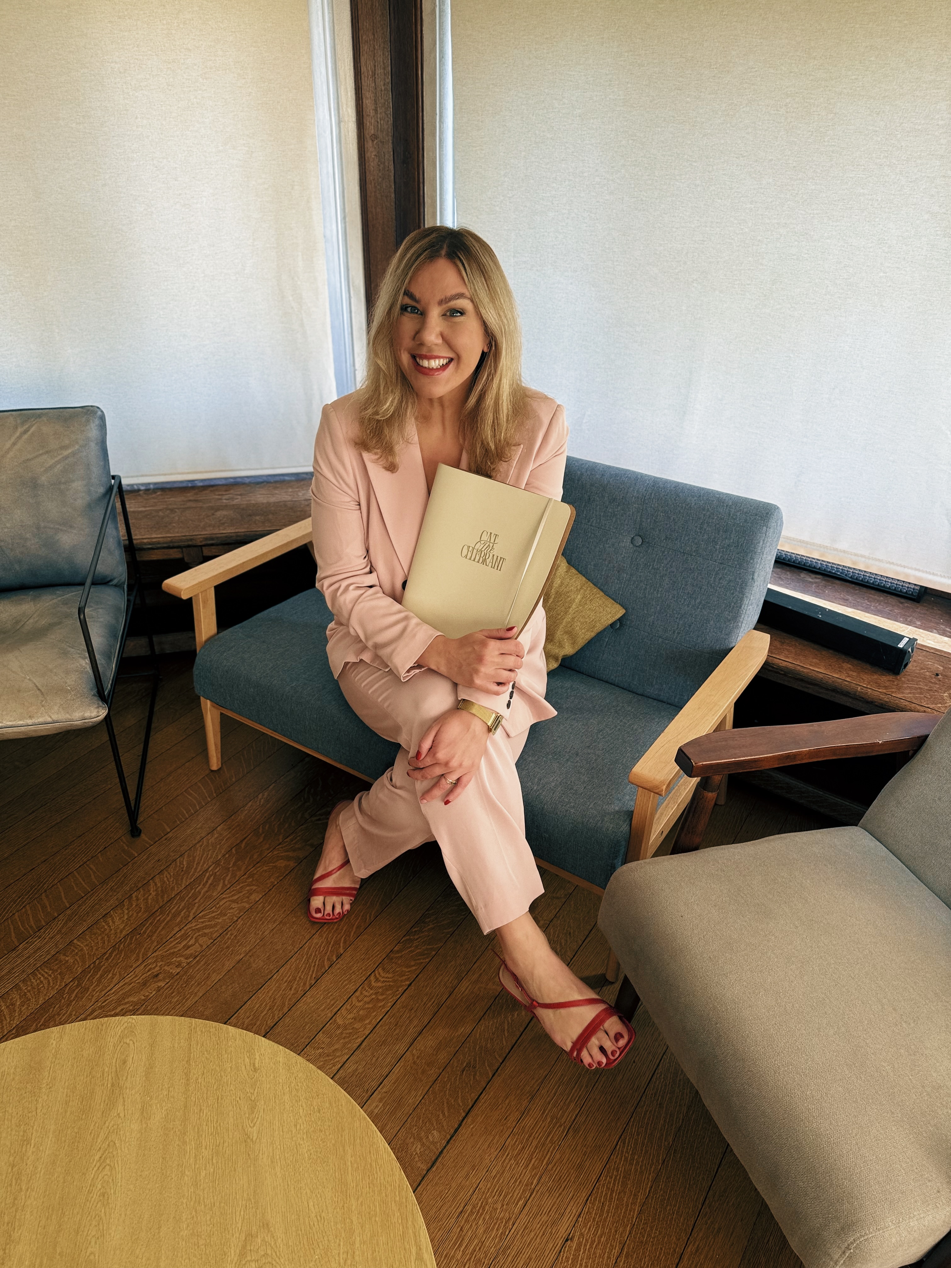 Cat the Celebrant, Queer Inclusive Celebrant in North London sits on a sofa in a pale pink suit holding her ceremony folder.