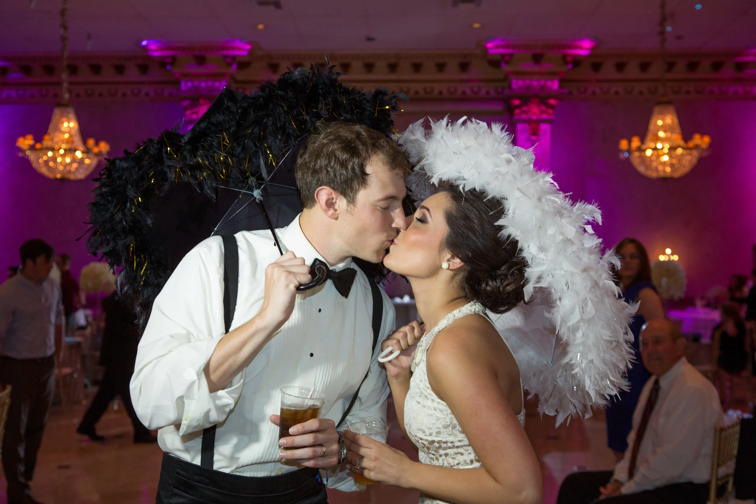 Bride and groom kissing indoors, each holding a feathered umbrella, with chandelier-lit background.